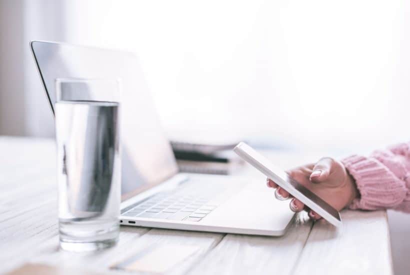 close up of white desk laptop clear glass of water with woman's hands holding phone