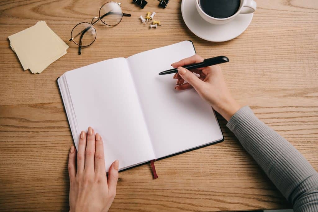 woman writing in journal with coffee and glasses on desk