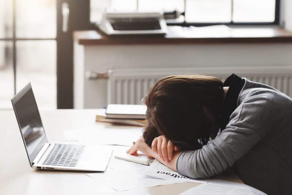 woman with head on desk with laptop and books